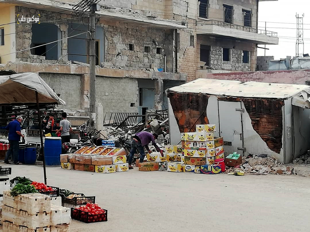 بسطات في سوق الأتارب وتظهر خلفها البيوت المدمرة بفعل القصف