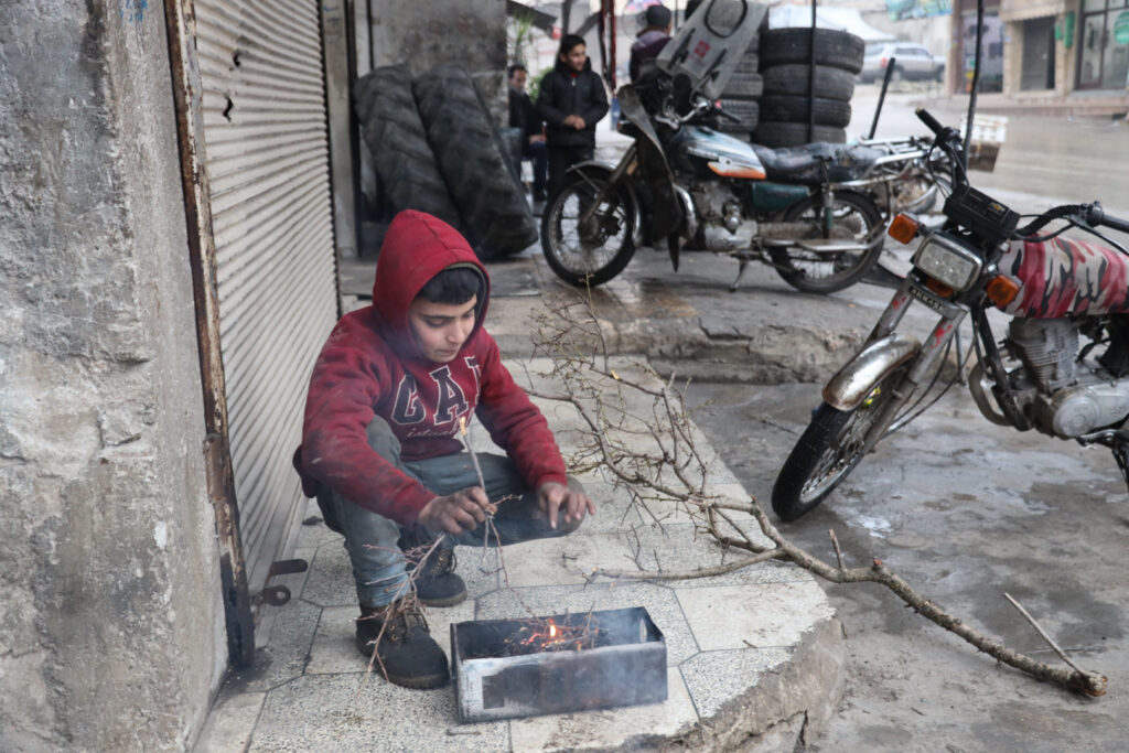طفل يحاول إشعال نار للتدفئة أمام محل الحدادة الذي يعمل به، مدينة كفرتخاريم غربي إدلب، شباط 2025- تصوير: رهام علي.