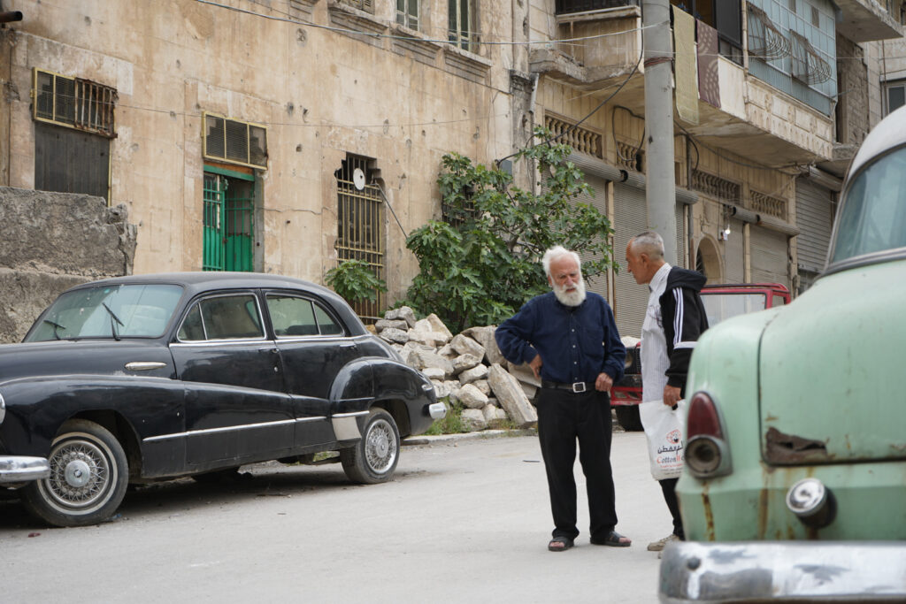 أبو عمر يقف بين سياراته المركونة أمام منزله، يشرح لرجل آخر عن السيارات وتاريخها، حي الشعار في مدينة حلب، 2025- تصوير: سحر زعتور.