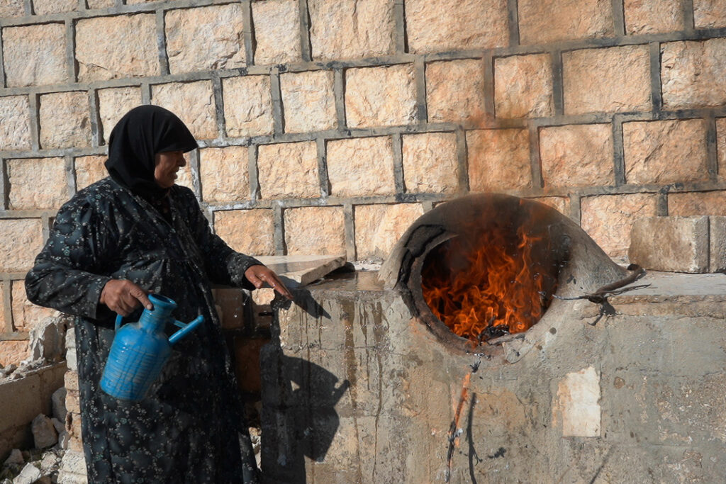 أم محمد تشعل تنور الجيران لإعداد الخبز، 
كفرنبل، ريف إدلب، أيار 2025- تصوير: سحر زعتور
