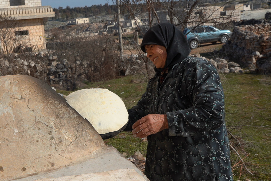 أم محمد تخبز على تنور جيرانها بعد أن هدمت الحرب التنور في منزلها، كفرنبل، ريف إدلب، أيار 2025- تصوير: سحر زعتور
