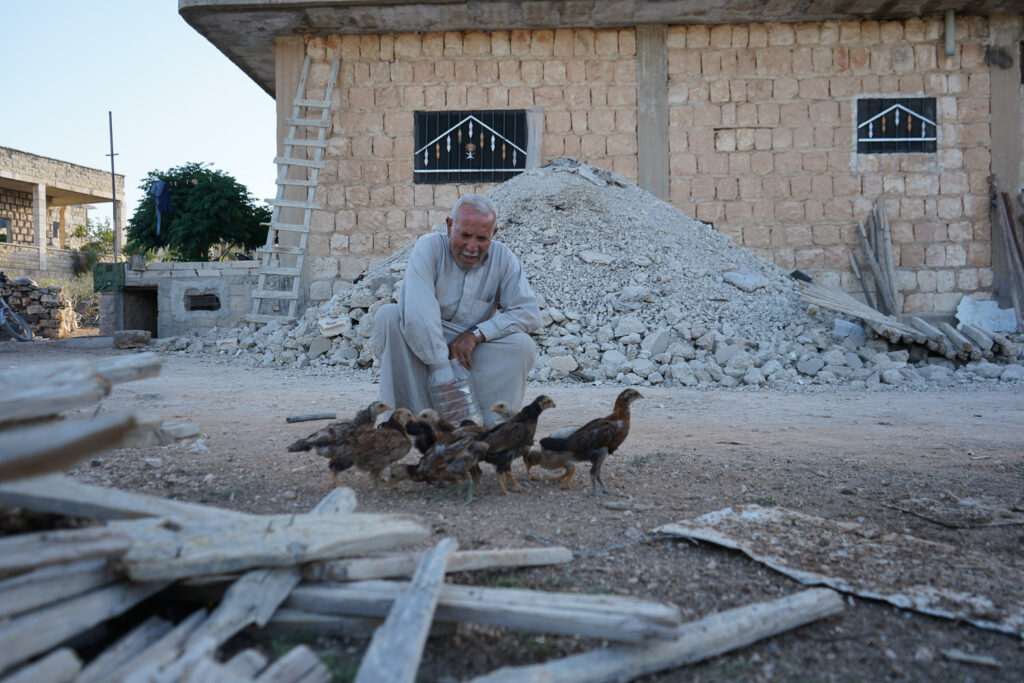 أبو محمد ينثر البذور للدجاجات لإطعامها، كفرنبل، ريف إدلب، أيار 2025- تصوير: سحر زعتور
