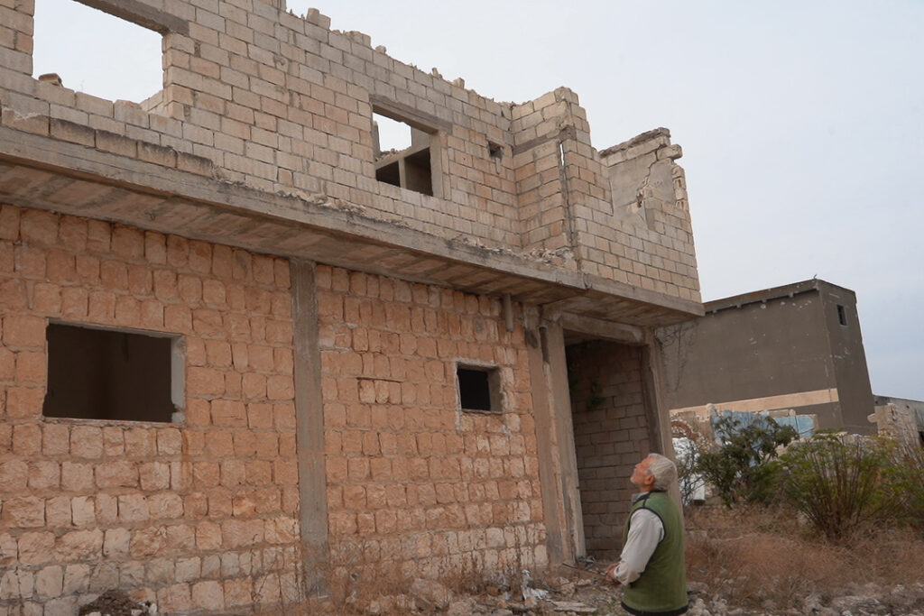 أبو محمد ينظر لمنزله الذي دمر قسم منه ونهبت أبوابه ونوافذه، كفرنبل، ريف إدلب، أيار 2025- تصوير: سحر زعتور
