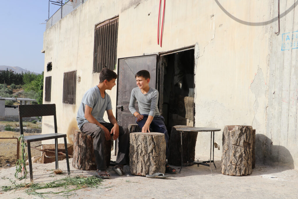 أحمد وعدنان ينتظران انتهاء مربي الأبقار من حلب أبقاره.  قرية مشمشان، جسر الشغور، أيار 2025- تصوير: هيام الباشا 