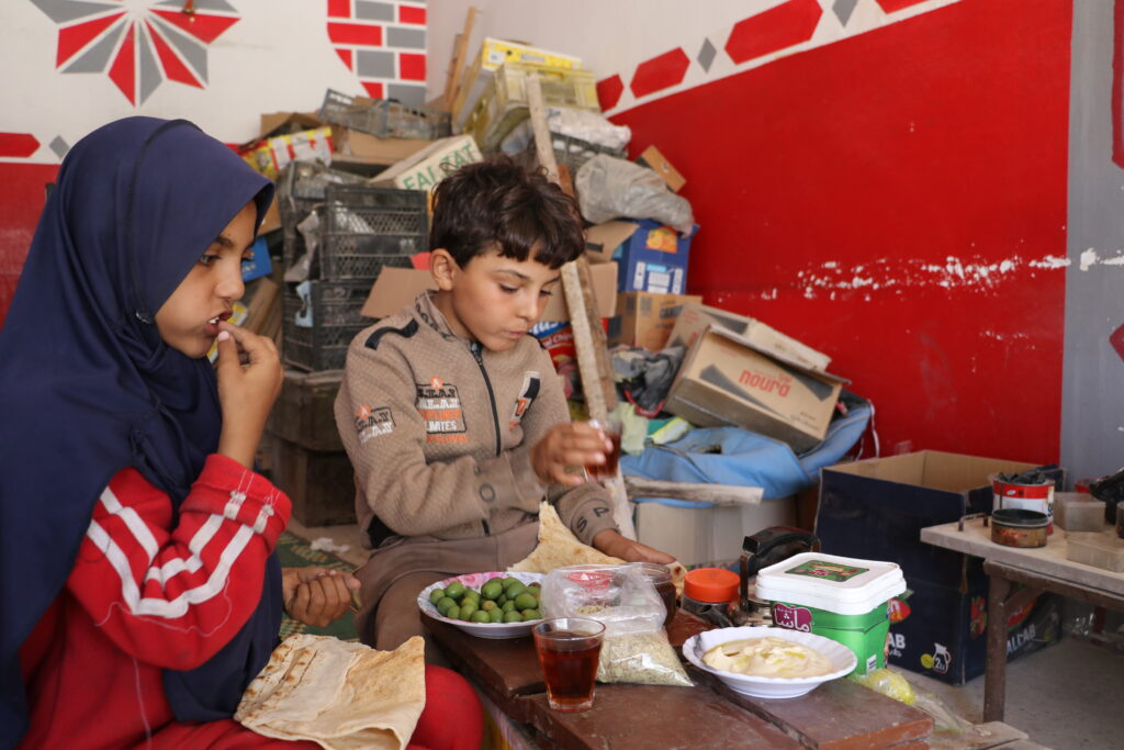  لميس ومحمد يتناولان طعام الغداء في محل جدهما قبل ذهابهما للبيت. خربة الجوز، ريف جسر الشغور. تشرين الأول 2025. تصوير: هيام الباشا